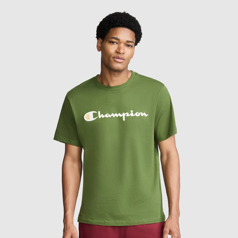 A man wearing the Champion Classic Graphic T-Shirt, Script Logo in green—with "Champion" displayed across the chest in bold white letters—stands against a plain light gray background, embodying timeless streetwear style.