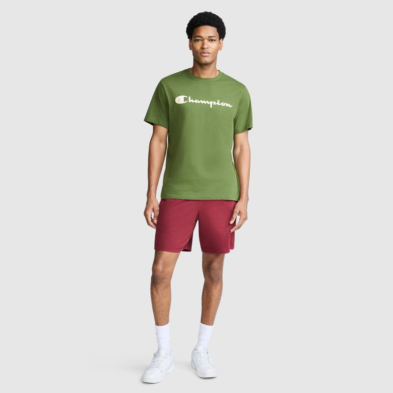 A person stands against a light gray background, wearing the Champion Classic Graphic T-Shirt with Script Logo styled as classic streetwear, paired with red shorts, white socks, and white sneakers.