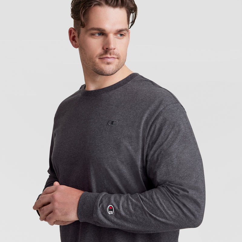 A man with short brown hair wears a Champion Classic Long Sleeve T-Shirt, Embroidered C Logo - Big & Tall, in dark gray. He stands against a plain light gray background, looking to his left with a neutral expression.