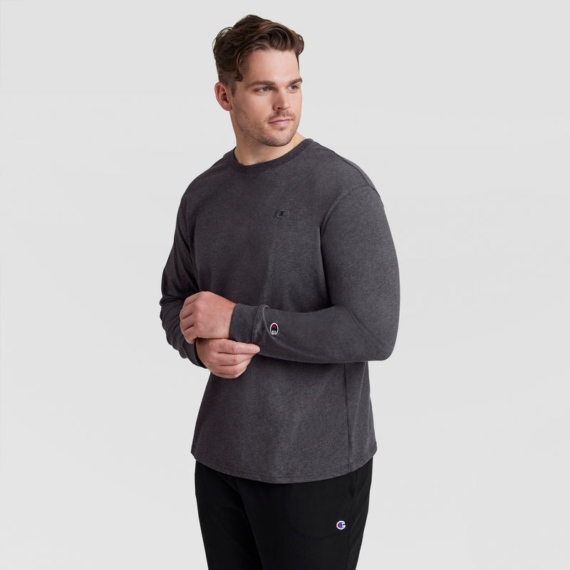 A man in a Champion Classic Long Sleeve T-Shirt with Embroidered C Logo (Big & Tall) stands against a light gray background, facing slightly to the side with his hands clasped in front.