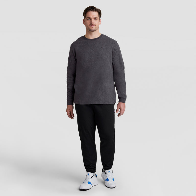 A man faces the camera against a light gray background, wearing a Champion Classic Long Sleeve T-Shirt with Embroidered C Logo (Big & Tall), black pants, and white sneakers with blue accents. He has a neutral expression.