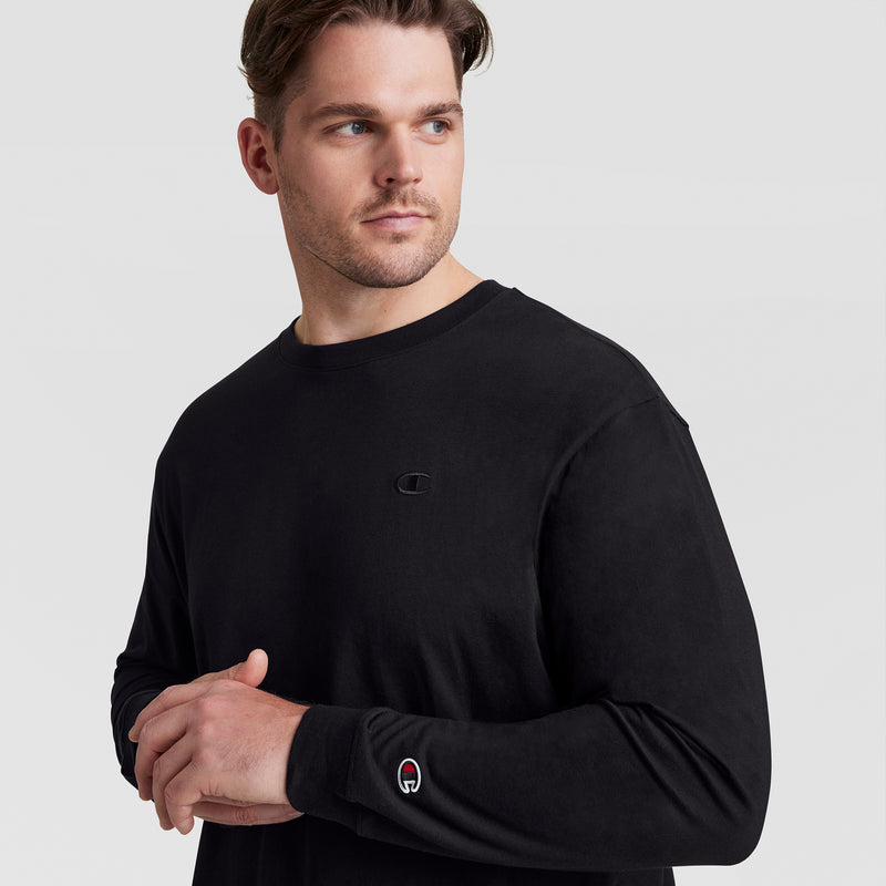 A man with short brown hair looks left against a plain light background, wearing the Champion Classic Long Sleeve T-Shirt, Embroidered C Logo - Big & Tall, featuring a small chest logo and embroidered sleeve patch.