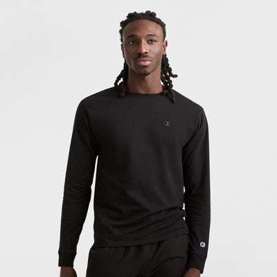 A man with long braided hair faces forward, wearing a Champion Classic Long Sleeve T-Shirt with Embroidered C Logo and black pants, set against a plain white background.