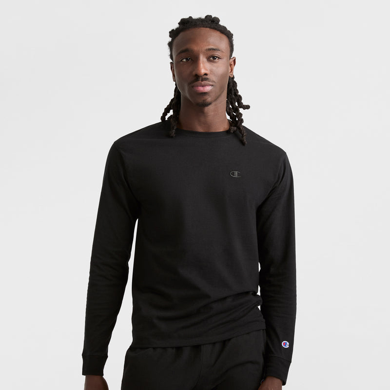 A man with long braided hair faces forward, wearing a Champion Classic Long Sleeve T-Shirt with Embroidered C Logo and black pants, set against a plain white background.