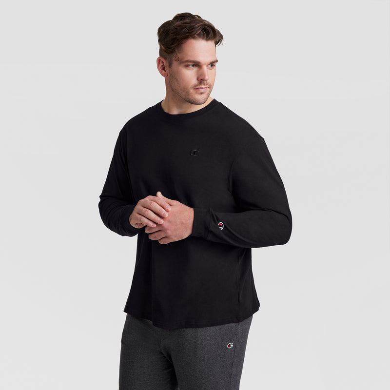 A man wearing the Champion Classic Long Sleeve T-Shirt with Embroidered C Logo (Big & Tall) in black and dark gray pants stands against a plain light gray background, looking to his left with his hands lightly clasped.
