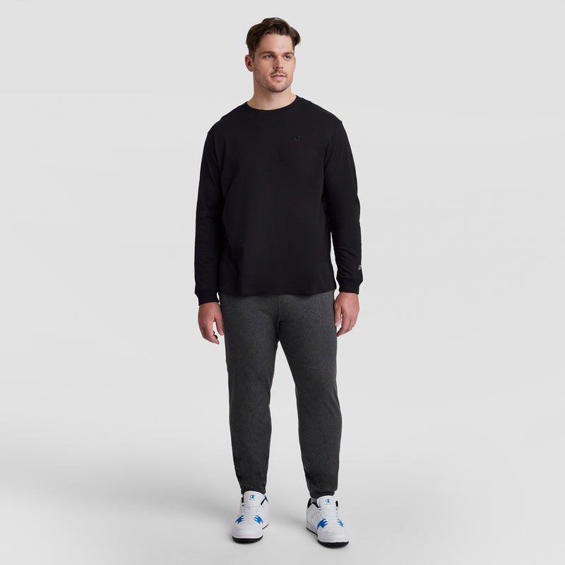 A man faces forward against a plain light gray background, wearing a black Champion Classic Long Sleeve T-Shirt with embroidered C logo (Big & Tall), dark gray jogger pants, and white sneakers with blue accents. He has a neutral expression.