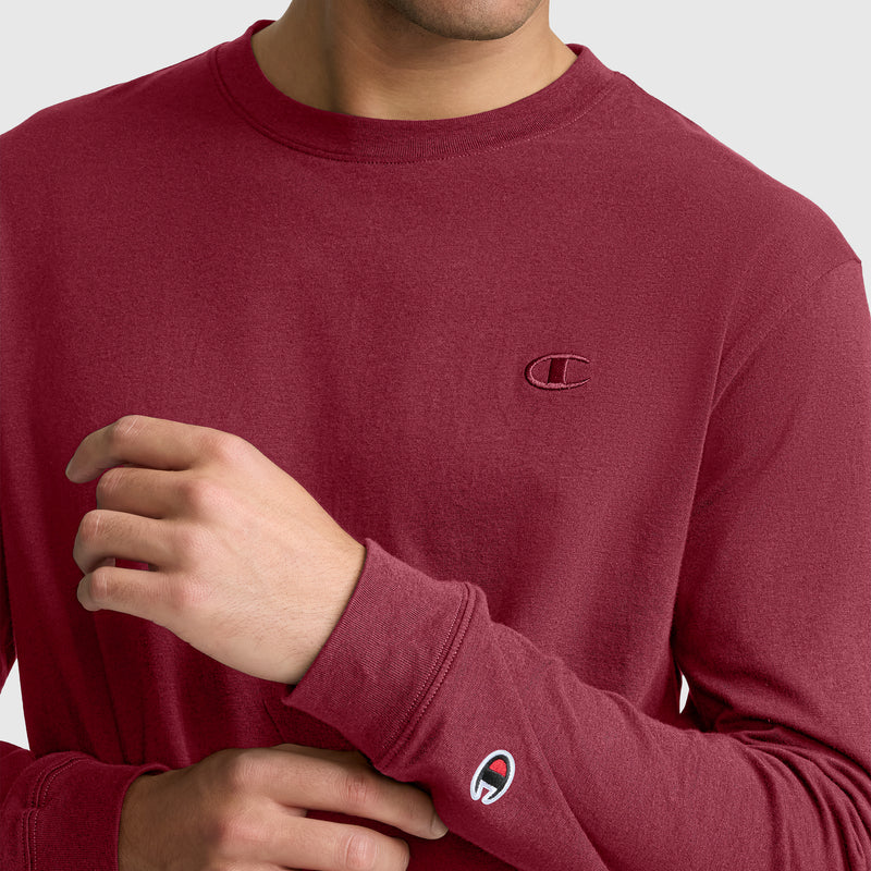 A person, partially out of frame, adjusts the sleeve of a maroon Champion Classic Long Sleeve T-Shirt with an embroidered C logo. This men’s shirt features the iconic Champion branding on the chest and wrist and is made from a comfortable cotton blend.