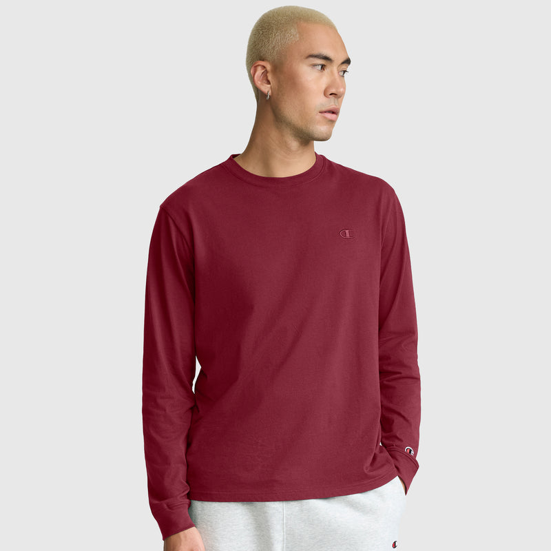 A person with short, blond hair stands against a plain light gray background, looking slightly to the side, wearing a maroon Champion Classic Long Sleeve T-Shirt with an embroidered C logo and light gray sweatpants.