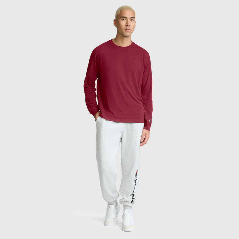 A person with short blond hair wears a Champion Classic Long Sleeve T-Shirt with an embroidered C logo in maroon, paired with light gray cotton blend sweatpants featuring a logo and white sneakers, standing against a plain light gray background.