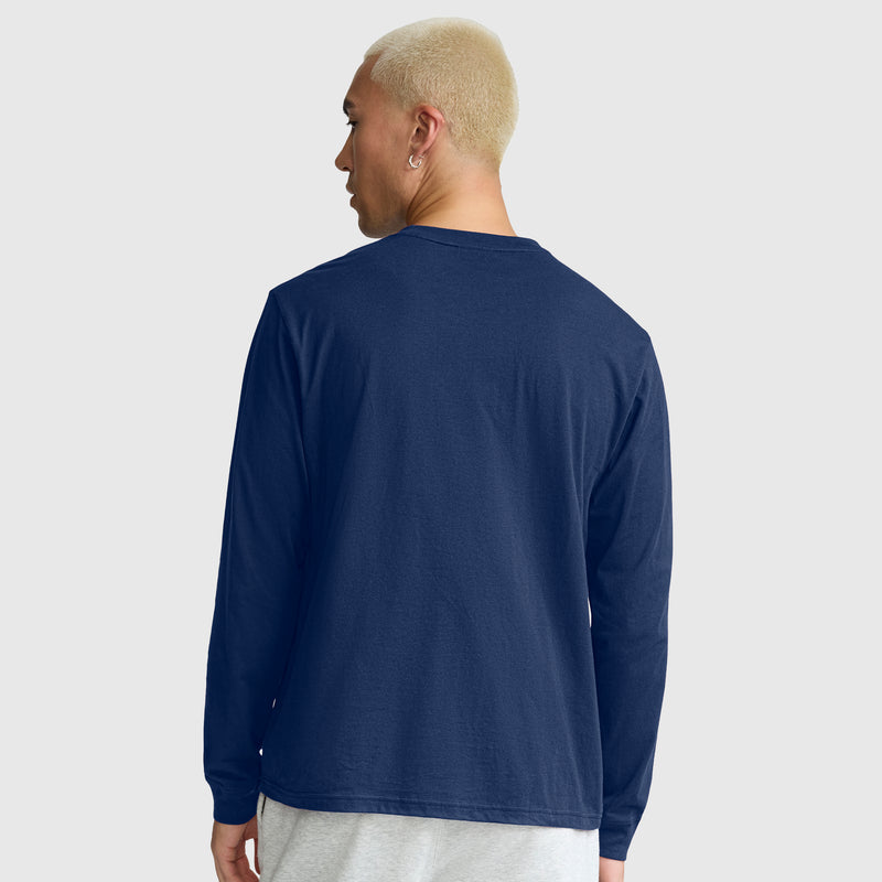A person with short blond hair is shown from the back against a plain light gray background, wearing light gray pants and a navy blue Champion Classic Long Sleeve T-Shirt featuring an embroidered C logo, crafted from a soft cotton blend.