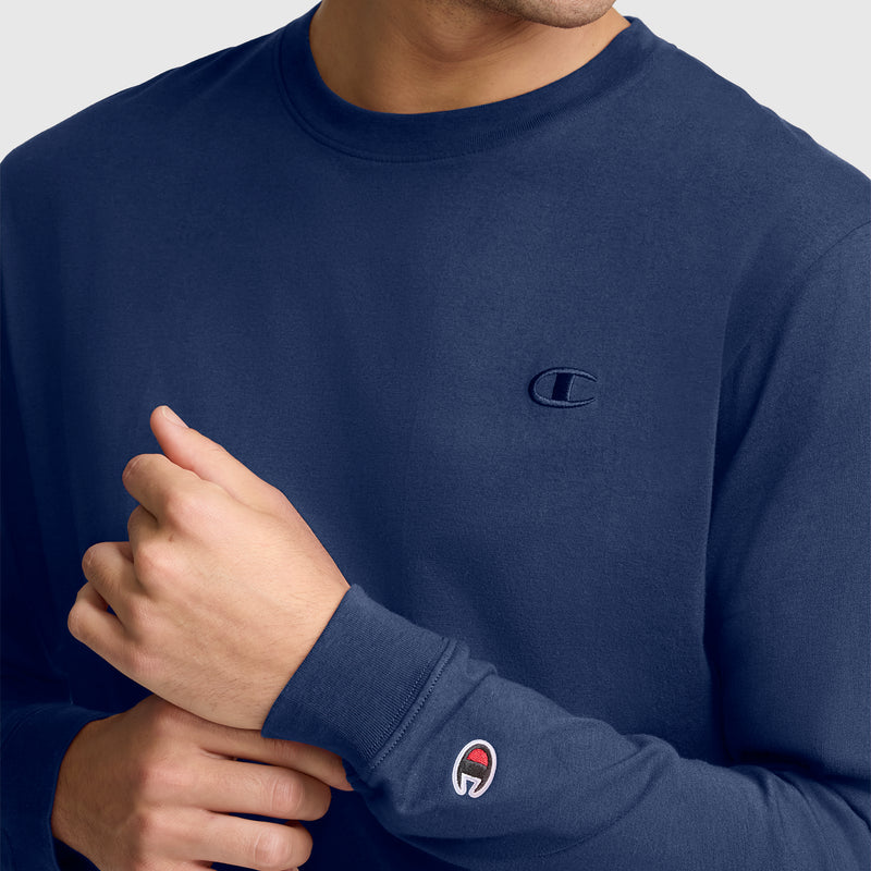 A person is wearing the Champion Classic Long Sleeve T-Shirt, Embroidered C Logo in navy blue. The iconic embroidered Champion “C” logo is visible on both the chest and sleeve. Made from a comfortable cotton blend, this casual athletic shirt fits well, with the wearer’s face partially out of frame.