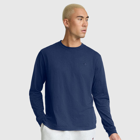 A person with short blond hair wears a navy blue Champion Classic Long Sleeve T-Shirt with an embroidered C logo, made from a soft cotton blend, along with light gray sweatpants. They stand against a plain light gray background, one hand in their pocket and looking to the side.