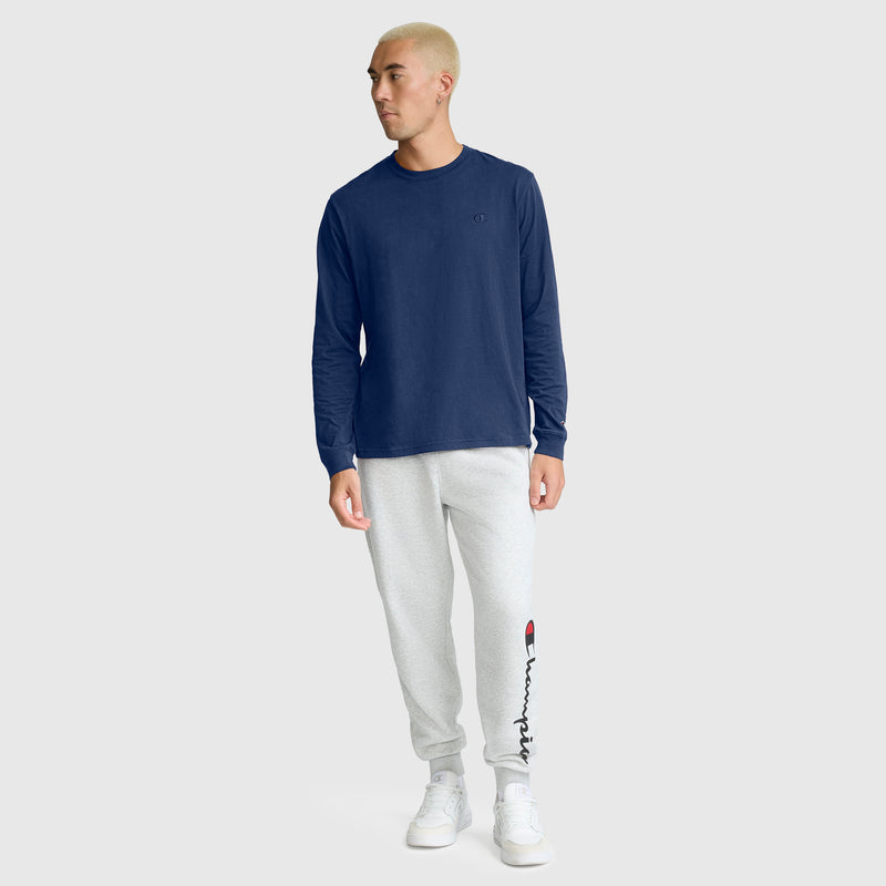 A person with short blond hair wears a Champion Classic Long Sleeve T-Shirt with Embroidered C Logo in navy, light gray jogger sweatpants featuring a logo on the leg, and white sneakers, standing against a plain light gray background.