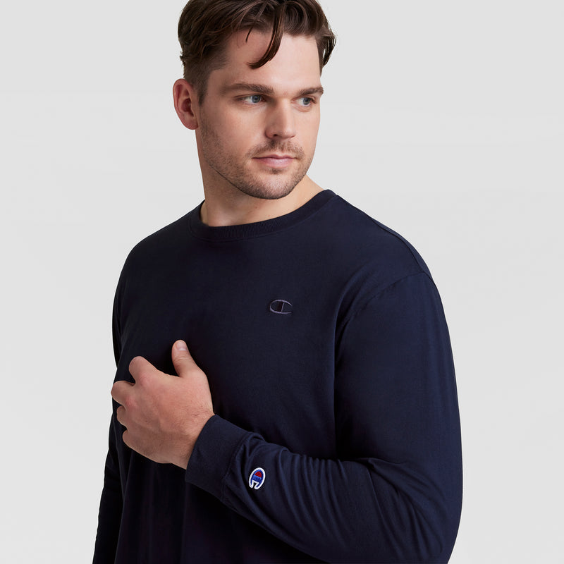 A man with short brown hair wears a Champion Classic Long Sleeve T-Shirt, Embroidered C Logo - Big & Tall in dark navy. He gazes to the side, left hand on his chest, against a plain light background.