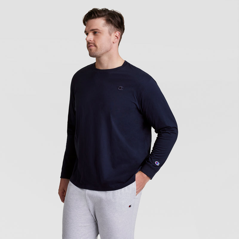 A man in a navy Champion Classic Long Sleeve T-Shirt, Embroidered C Logo - Big & Tall, and light gray sweatpants stands against a plain background, looking left with hands in pockets for a sporty, relaxed vibe.