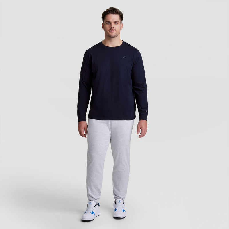 A man stands facing forward wearing a navy Champion Classic Long Sleeve T-Shirt with an embroidered C logo (Big & Tall), light gray sweatpants, and white sneakers with blue accents, against a plain light gray background.