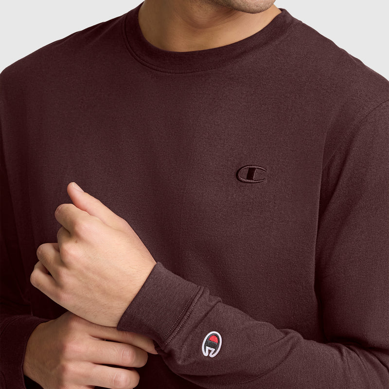 A person is shown wearing the Champion Classic Long Sleeve T-Shirt with Embroidered C Logo in maroon, featuring visible chest and arm logos. The image focuses on the lower face and torso, with one hand adjusting the sleeve of this comfortable men’s athletic shirt crafted from a soft cotton blend.