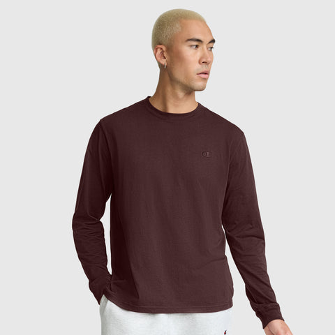 A person with short blond hair wears a maroon Champion Classic Long Sleeve T-Shirt, Embroidered C Logo, paired with light gray pants, standing with one hand in a pocket against a plain light gray background.