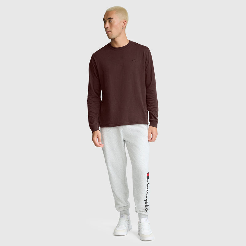 A person with short blond hair wears a maroon Champion Classic Long Sleeve T-Shirt with an embroidered C logo, light gray sweatpants featuring a logo on the left leg, and white sneakers while standing against a plain light gray background.