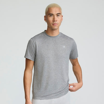A person with short, bleached blonde hair wears a grey short-sleeve t-shirt and light pants, standing against a plain light background while gently pulling the hem of their shirt with one hand.