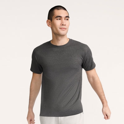 A man with short dark hair wears a Champion Classic T-Shirt, C Logo in dark gray and light gray pants, standing against a light background and looking slightly to his left with a neutral expression.