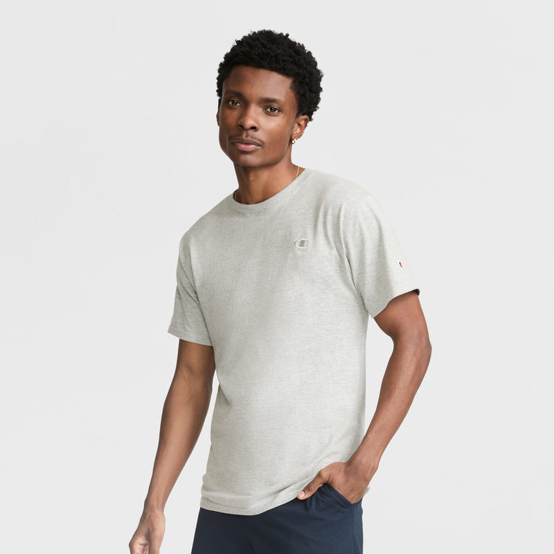 A man wearing a Champion Classic T-Shirt, C Logo in light gray and dark pants stands against a plain white background, looking at the camera with a neutral expression.