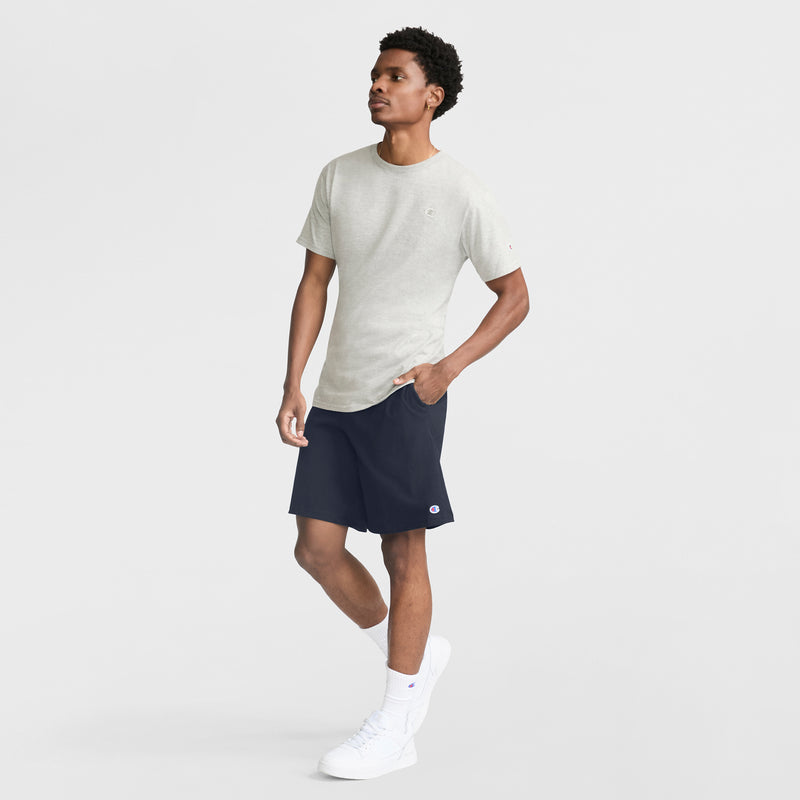 A man stands with one hand in his pocket against a plain white background, wearing a Champion Classic T-Shirt, C Logo in light gray, paired with dark navy shorts, white socks, and white sneakers.