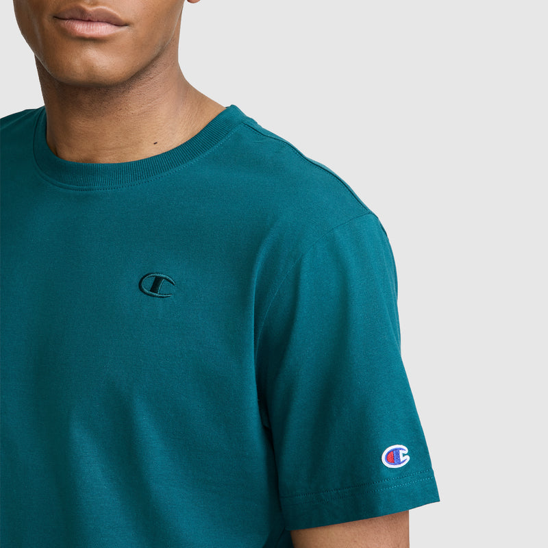 A person wearing a teal Champion Classic T-Shirt, C Logo, featuring the signature Champion branding on the chest and sleeve, stands against a light gray background.
