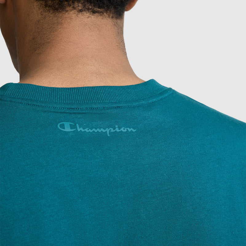 The back of a person's neck and shoulders wearing the Champion Classic T-Shirt, C Logo in teal cotton with "Champion" printed near the collar on the upper back, embodying the relaxed style of this iconic men's tee.