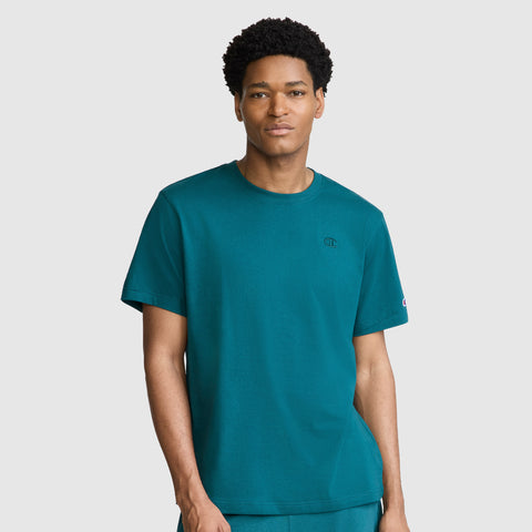 A man with short curly hair wears the Champion Classic T-Shirt, C Logo in teal, paired with matching pants, standing against a plain light gray background.