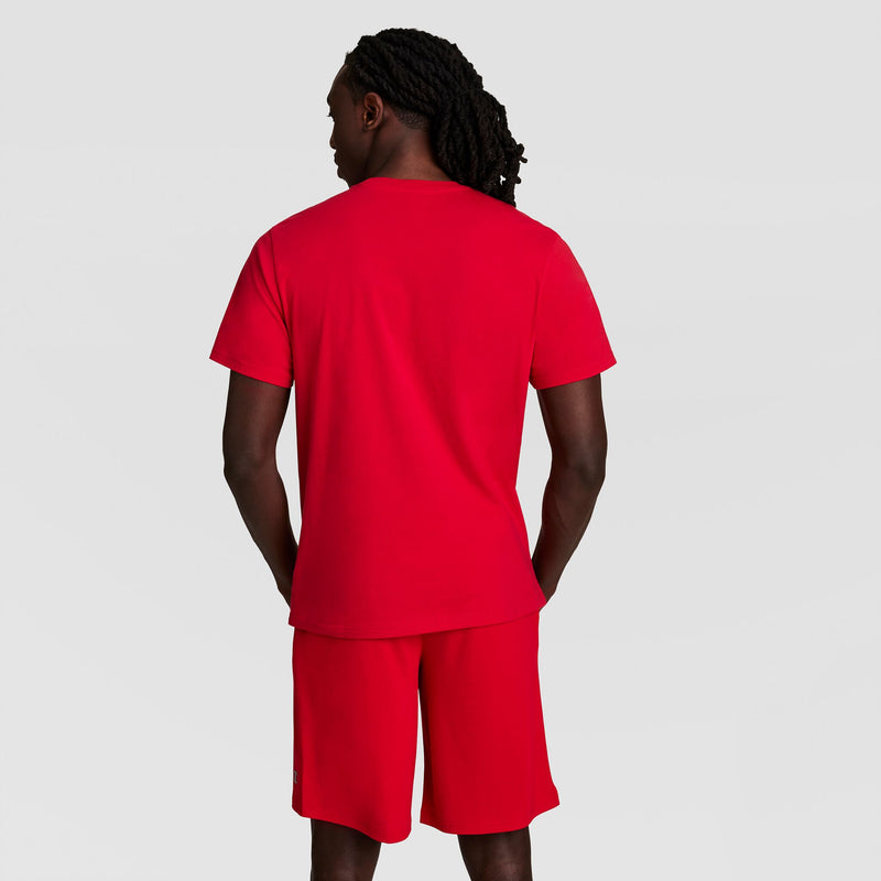 A man with long, braided hair faces away, wearing a matching red Champion Classic T-Shirt with C Logo and red shorts against a plain light gray background.