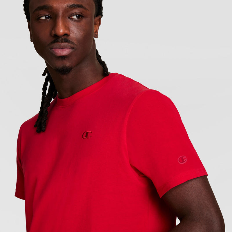 A man with long braids wears the Champion Classic T-Shirt, C Logo, in bright red, featuring small chest and sleeve logos. He looks to his left against a plain light gray background.