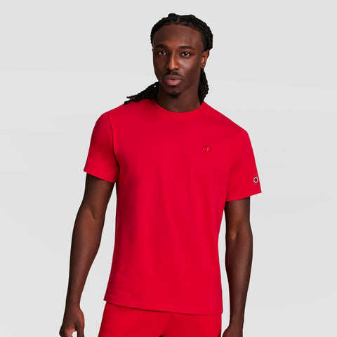 A man with long braided hair wears the Champion Classic T-Shirt with C Logo in bright red, paired with matching red pants, standing against a plain light gray background.