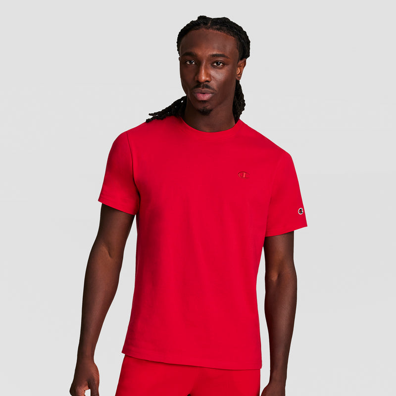 A man with long braided hair wears the Champion Classic T-Shirt with C Logo in bright red, paired with matching red pants, standing against a plain light gray background.