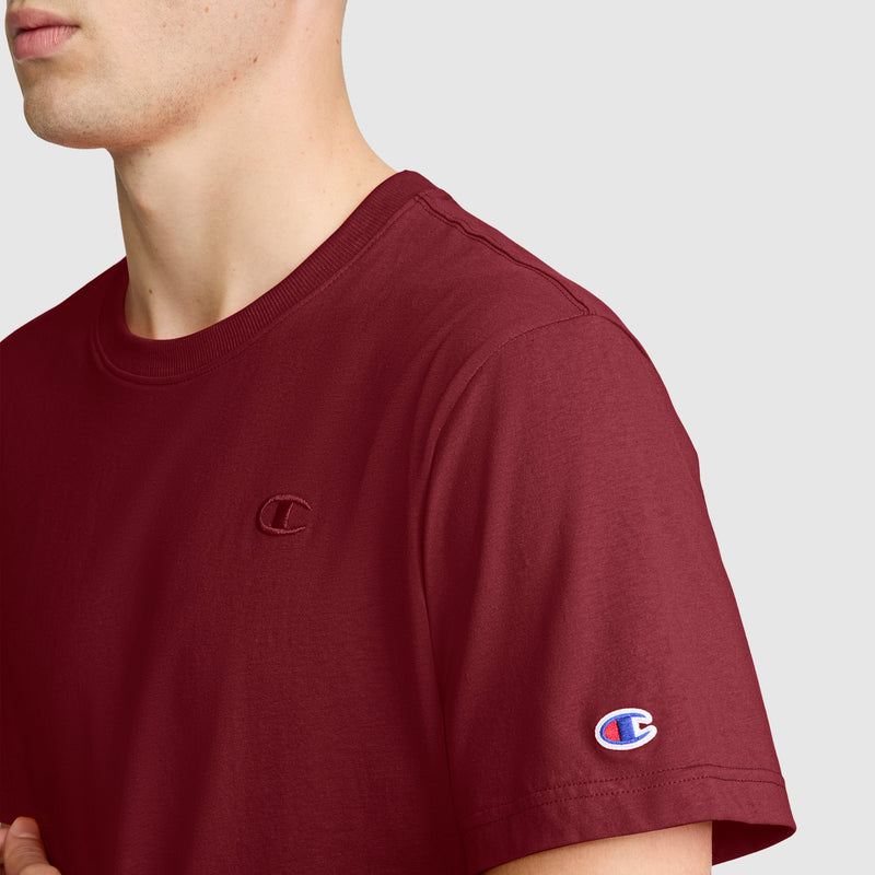 A person is shown from the shoulders up against a plain light background, wearing the Champion Classic T-Shirt, C Logo in maroon. This men’s tee features the brand’s signature embroidered C logo on the chest and sleeve, blending iconic Champion style with everyday comfort.