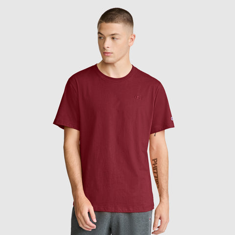 A young man wears the Champion Classic T-Shirt with C Logo in plain maroon, paired with gray shorts, standing against a light gray background and looking slightly left with a neutral expression.