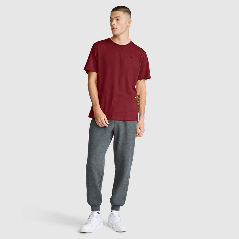 A person wearing the Champion Classic T-Shirt, C Logo in maroon, paired with gray jogger pants and white sneakers, stands against a plain light gray background, looking to the side.
