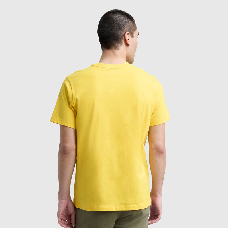 A person with short dark hair is standing with their back to the camera, wearing a yellow Champion Classic T-Shirt with a C Logo and olive green pants, against a light gray background.