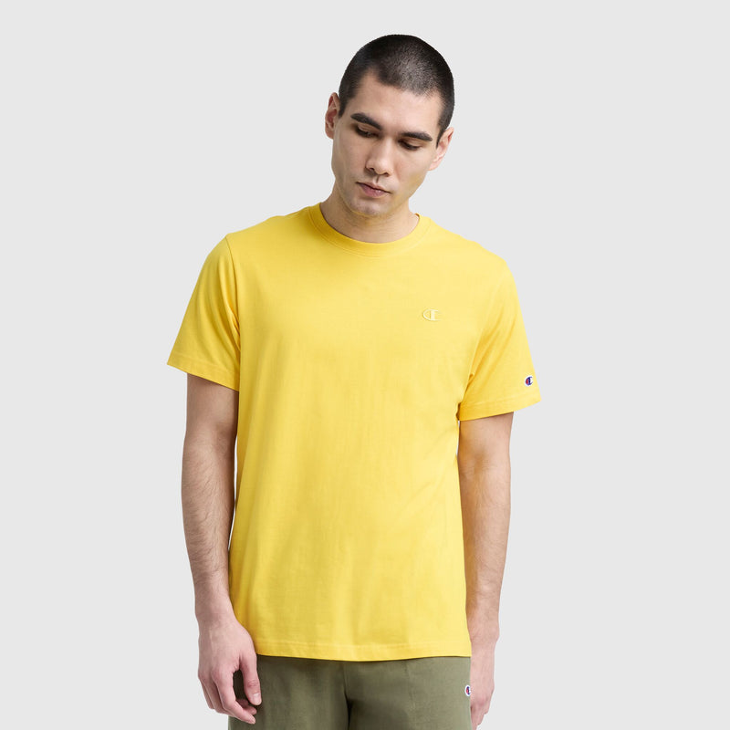A man with short dark hair wears a Champion Classic T-Shirt with C Logo in yellow and olive green pants, standing against a light gray background. He looks down, arms relaxed at his sides, embodying classic men's style.