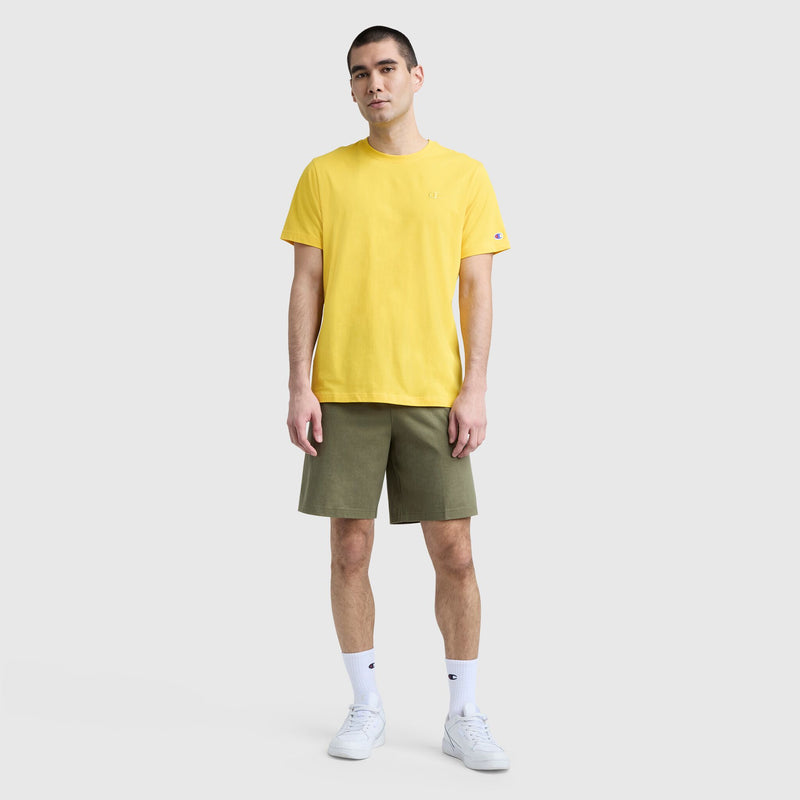 A man models classic men's style in front of a light gray backdrop, wearing a yellow Champion Classic T-Shirt with C Logo, olive green shorts, white socks, and white sneakers. He stands facing forward with his arms relaxed at his sides.