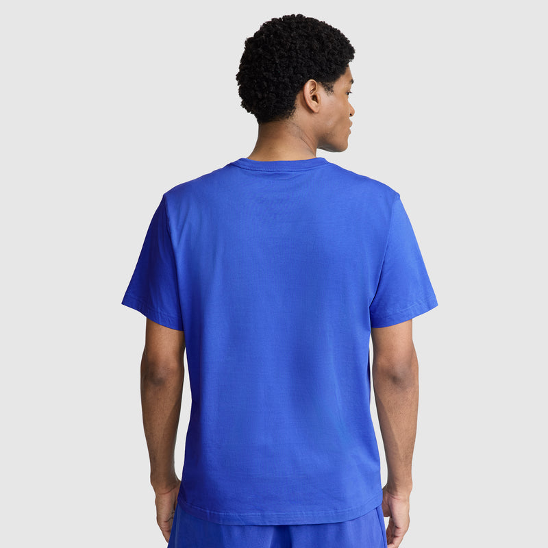 A person stands facing away from the camera, wearing a bright blue Classic T-Shirt with Champion’s C Logo and matching blue shorts, against a plain light gray background.