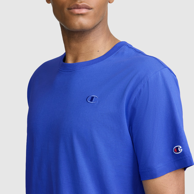 Someone is wearing Champion's Classic T-Shirt, featuring the iconic C logo on the chest and sleeve, in a vibrant blue color. The men's tee is crafted from soft cotton and shown against a neutral background.