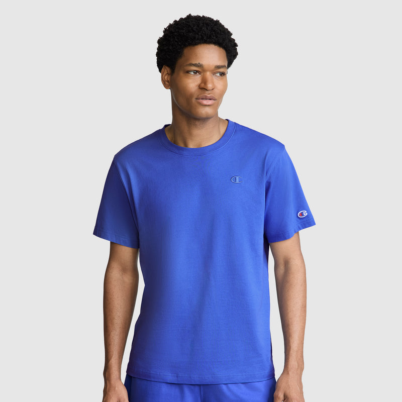 A man wearing a bright blue Champion Classic T-Shirt with the C Logo and matching shorts stands against a plain light gray background, looking slightly to his left.