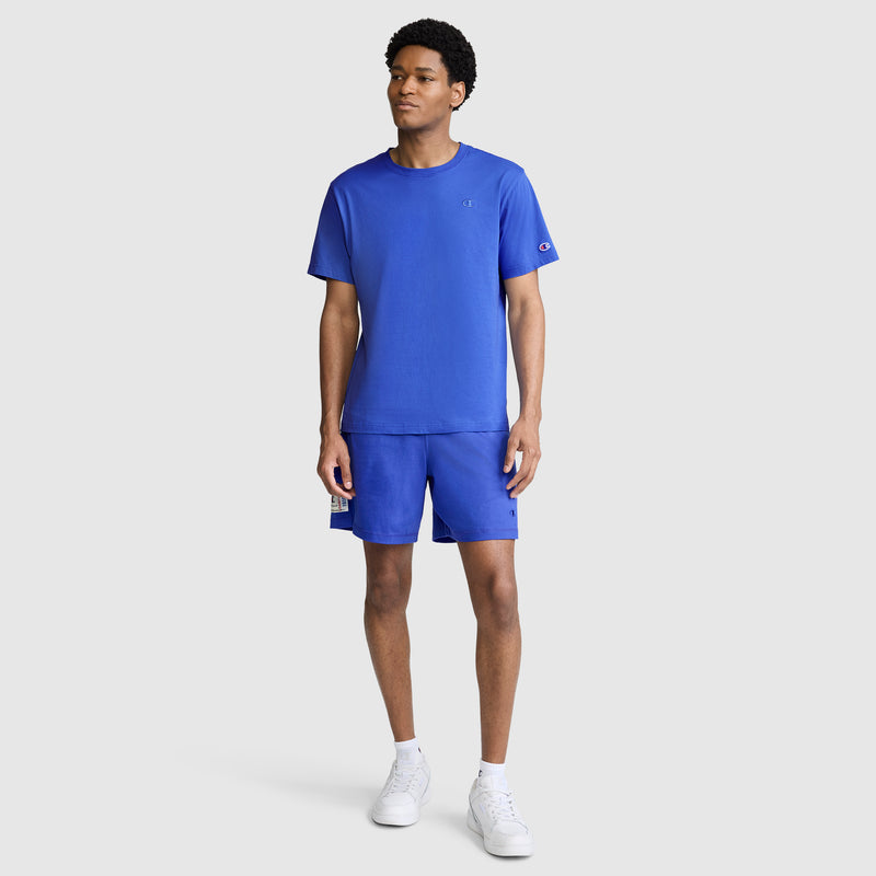 A man stands facing forward, wearing a matching blue Champion Classic T-Shirt, C Logo, and blue shorts. His look is completed with white socks and white sneakers against a plain light gray background.