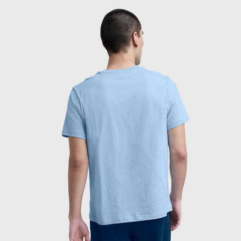 A person with short dark hair faces away, wearing a light blue Champion Classic T-Shirt with C Logo and dark blue pants against a plain light gray background.