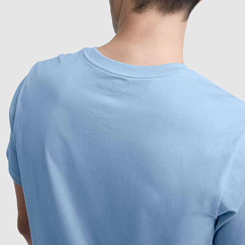 A person is shown from behind wearing the Champion Classic T-Shirt, C Logo in light blue cotton with short sleeves and subtle embroidery near the collar. The neck and shoulders are visible against a plain, light-colored background.