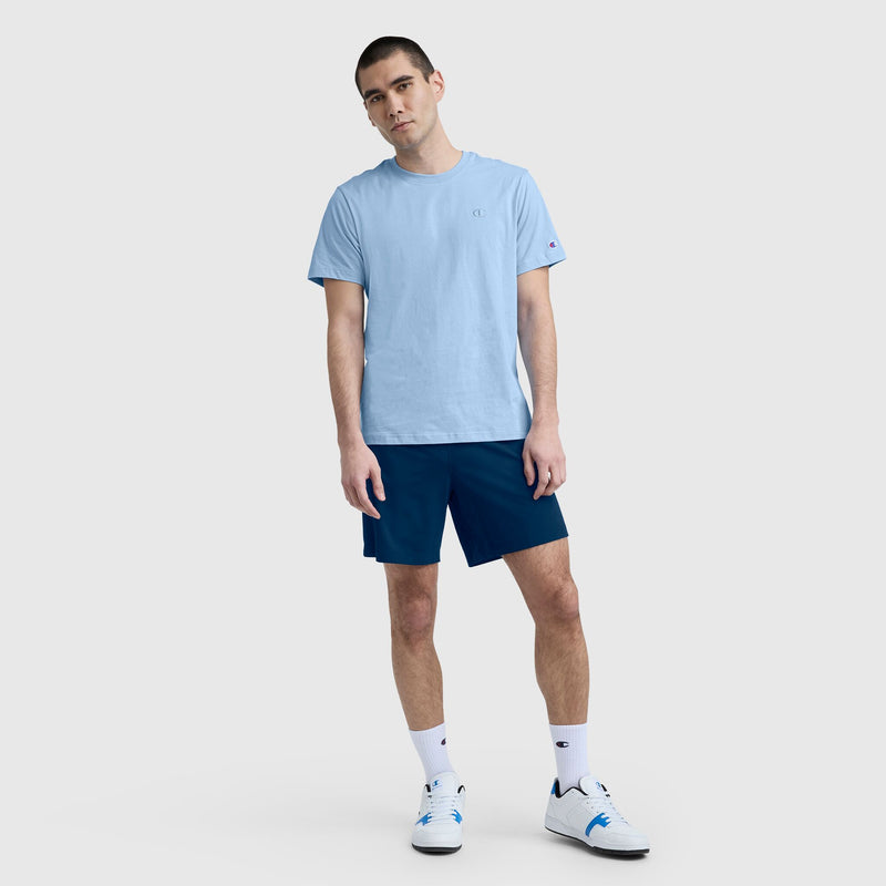 A man with short dark hair stands against a plain gray background, wearing a Champion Classic T-Shirt, C Logo in light blue, dark blue shorts, white socks, and white sneakers. He stands relaxed with his arms at his sides.
