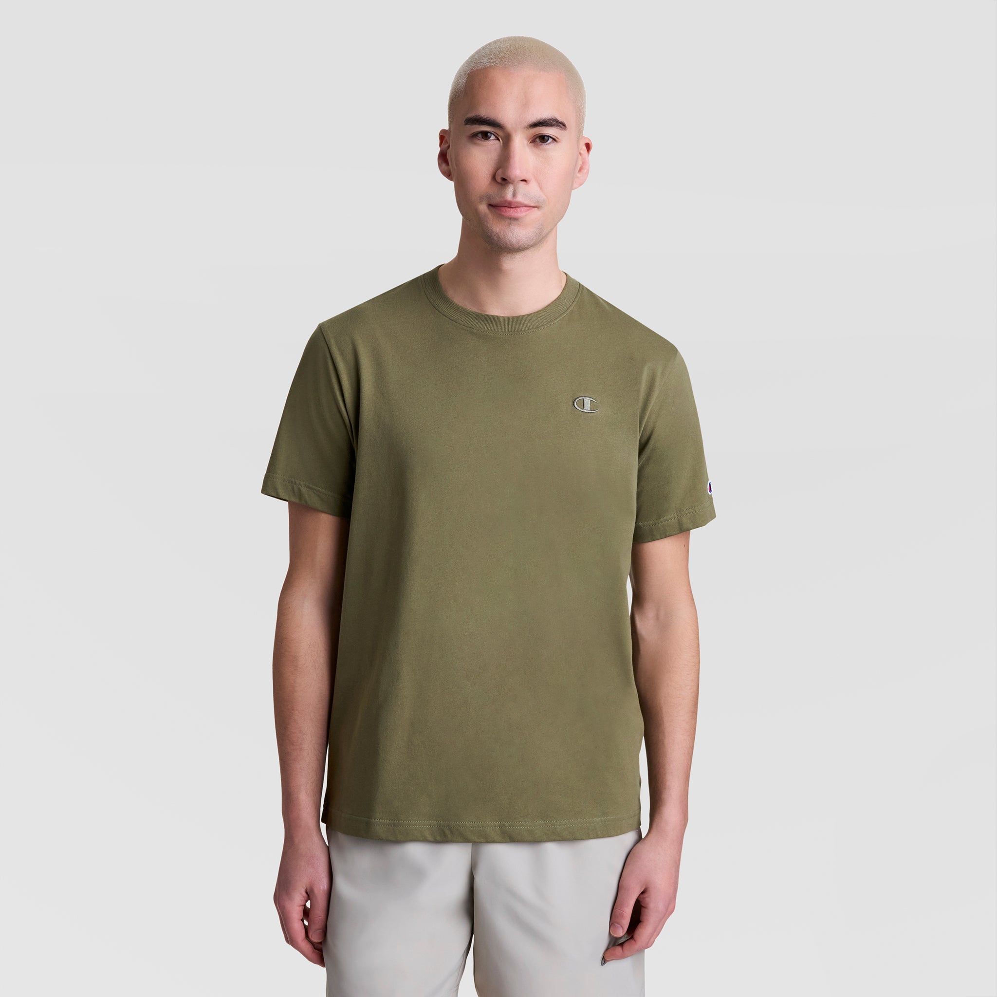 A person with short, light blonde hair is wearing a Champion Classic T-Shirt, C Logo in olive green and light gray shorts, facing the camera against a plain light background.