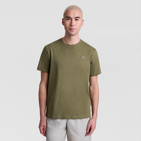 A person with short, light blonde hair is wearing a Champion Classic T-Shirt, C Logo in olive green and light gray shorts, facing the camera against a plain light background.