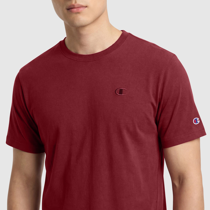 A person is shown from shoulders to mid-torso wearing Champion's Classic T-Shirt, C Logo, a plain maroon cotton tee with the logo embroidered on the chest and sleeve, set against a light background.